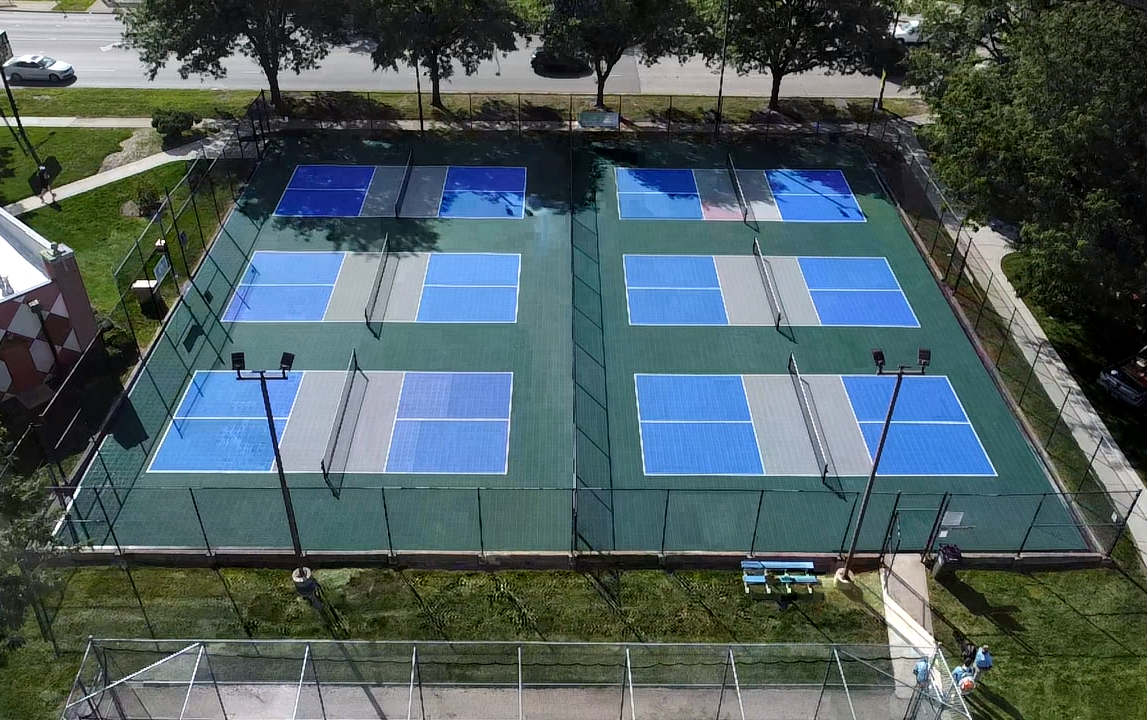 Aerial view of a tennis court with multiple blue and gray courts, surrounded by fencing and trees.