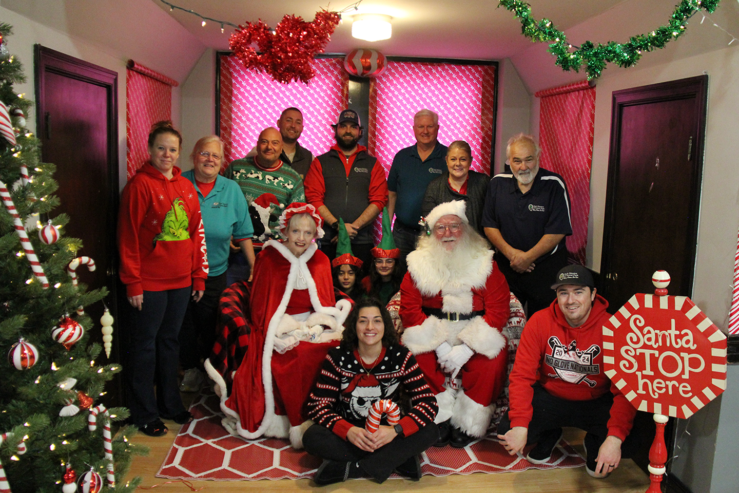 Group photo of people celebrating Christmas, including Santa Claus and a woman dressed as Mrs. Claus, all smiling and surrounded by Christmas decorations such as a tree with ornaments, candy canes, tinsel, and a red and white themed backdrop.