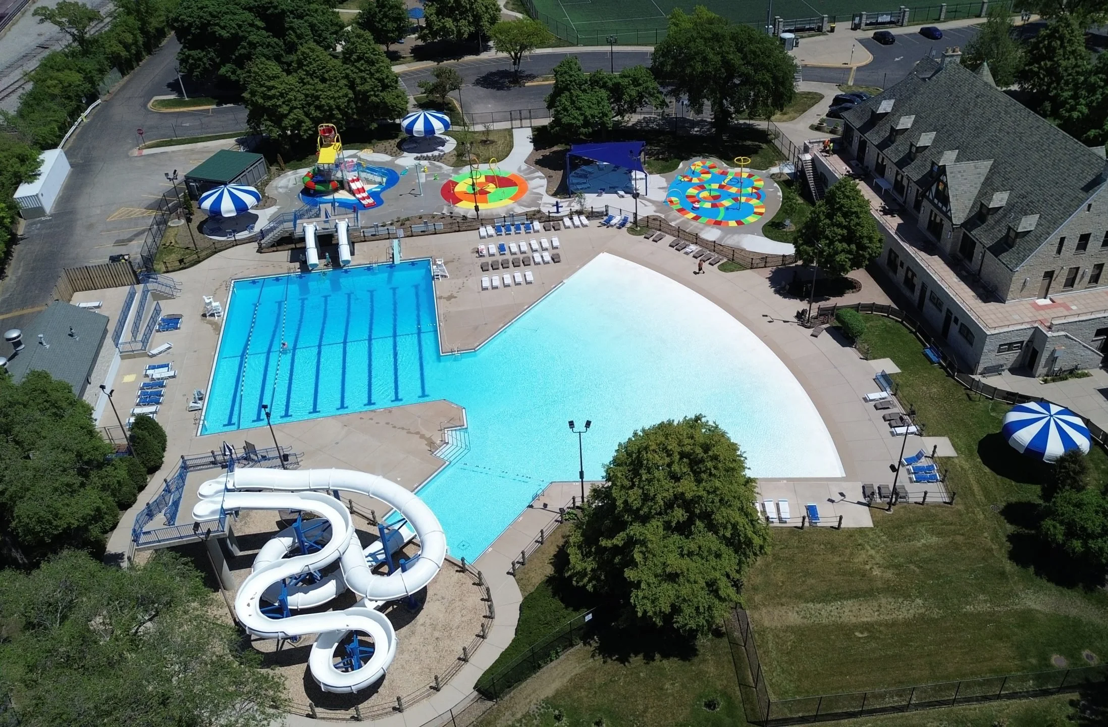 An aerial view of a colorful outdoor water park featuring a large swimming pool with water slides, a splash pad with colorful patterns, shaded seating areas, and trees surrounding the area.