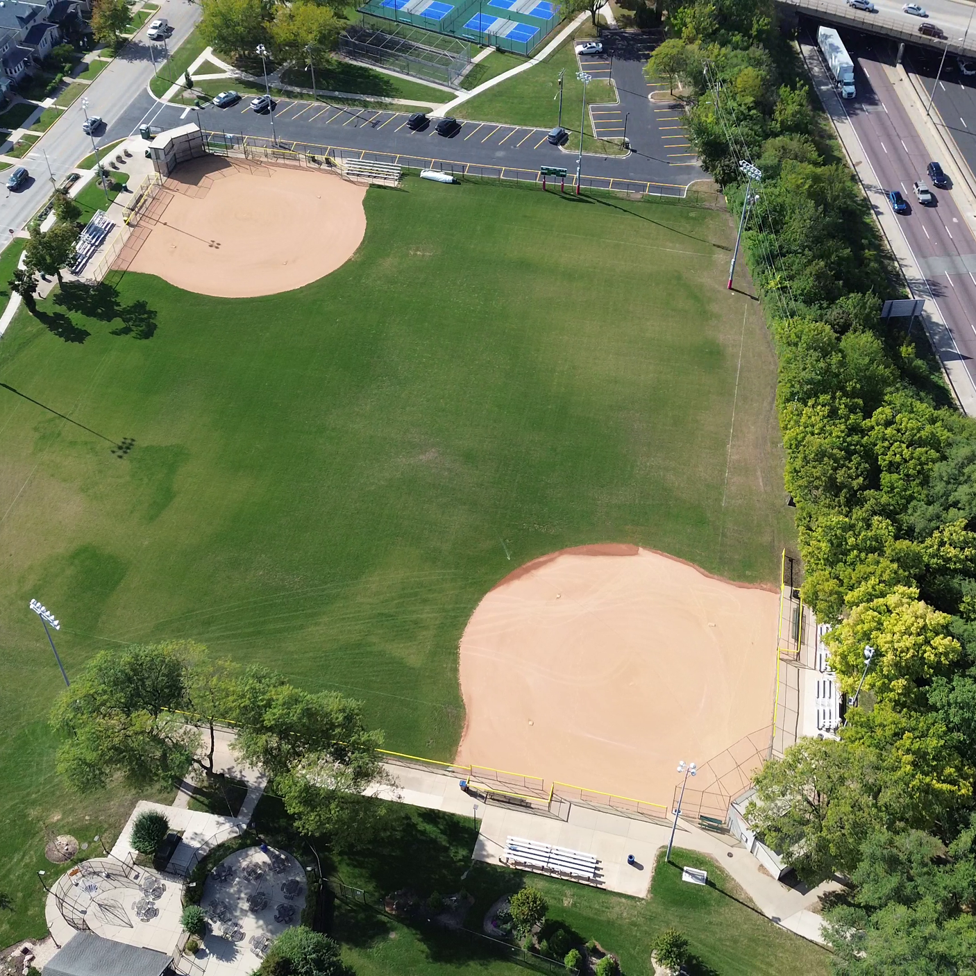 An aerial view of a baseball field with two playing areas, surrounded by trees, parking lots, and nearby roads.