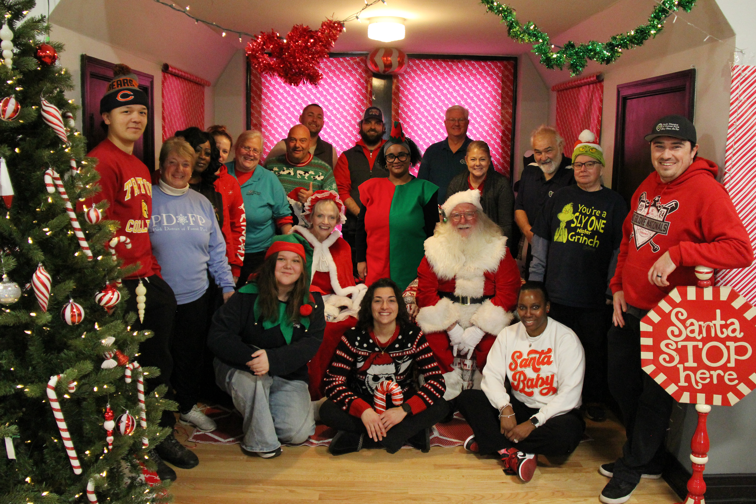 Group of people gathered around Santa Claus and Mrs. Claus, celebrating Christmas indoors. Decorated Christmas tree, holiday ornaments, and colorful decorations are visible.