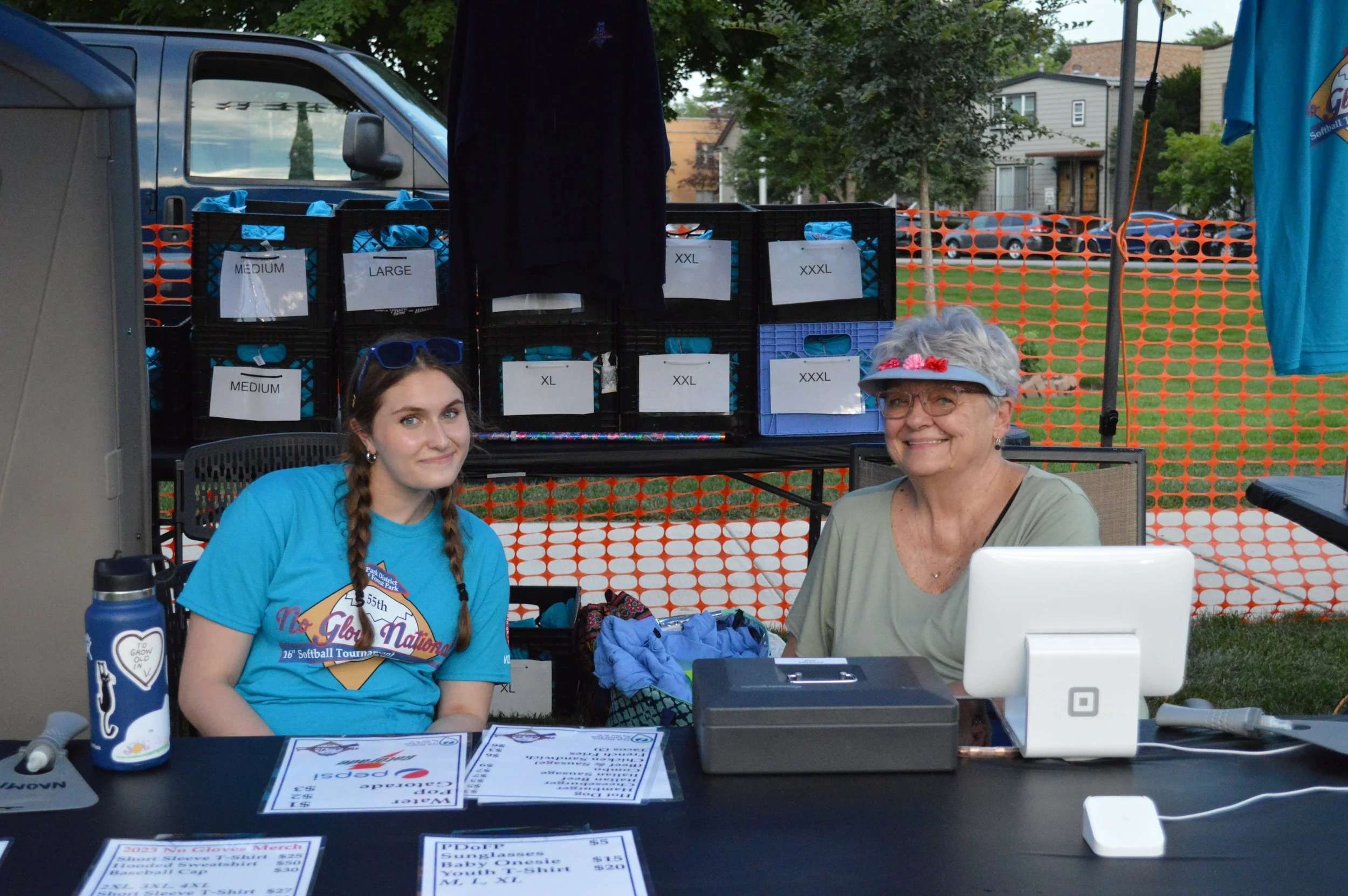 Two women sitting at a booth, one young with braided hair wearing sunglasses and a blue T-shirt, and an older woman with gray hair wearing a visor and glasses, with a costume contest sign and price list on the table, and boxes labeled with shirt sizes behind them.