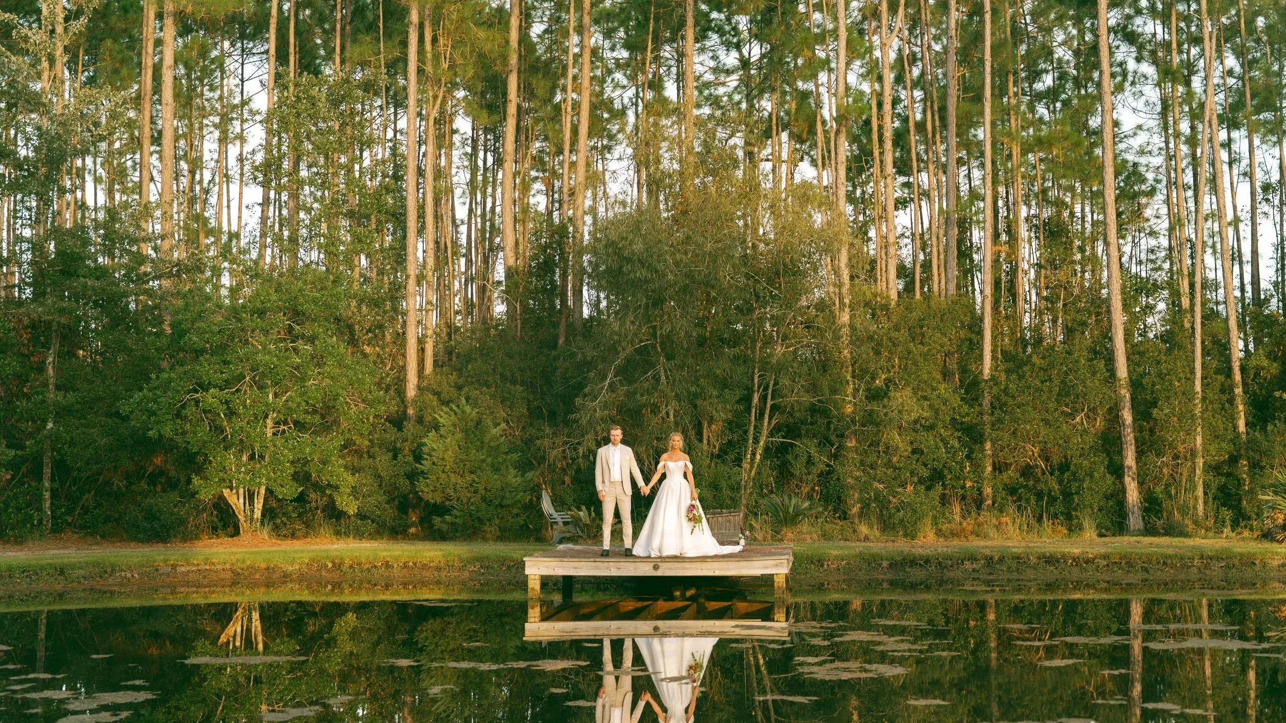A bride and groom in wedding attire standing on a wooden dock by a calm lake, holding hands, with a backdrop of tall pine trees and green foliage, reflected in the water.