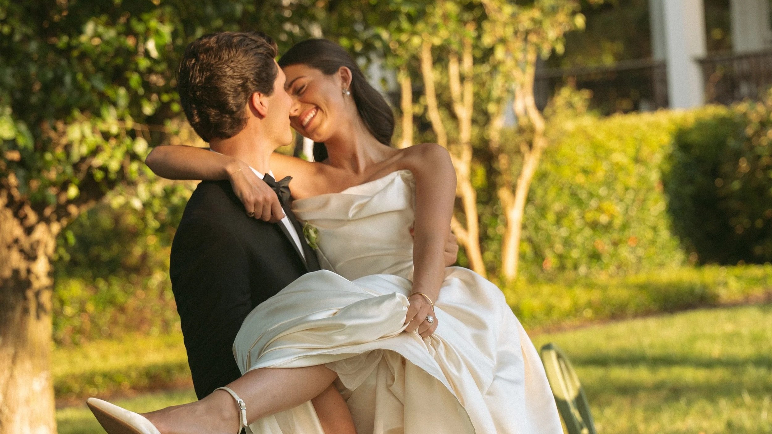Groom in a tuxedo lifting bride in a wedding dress outdoors with trees and greenery in the background.