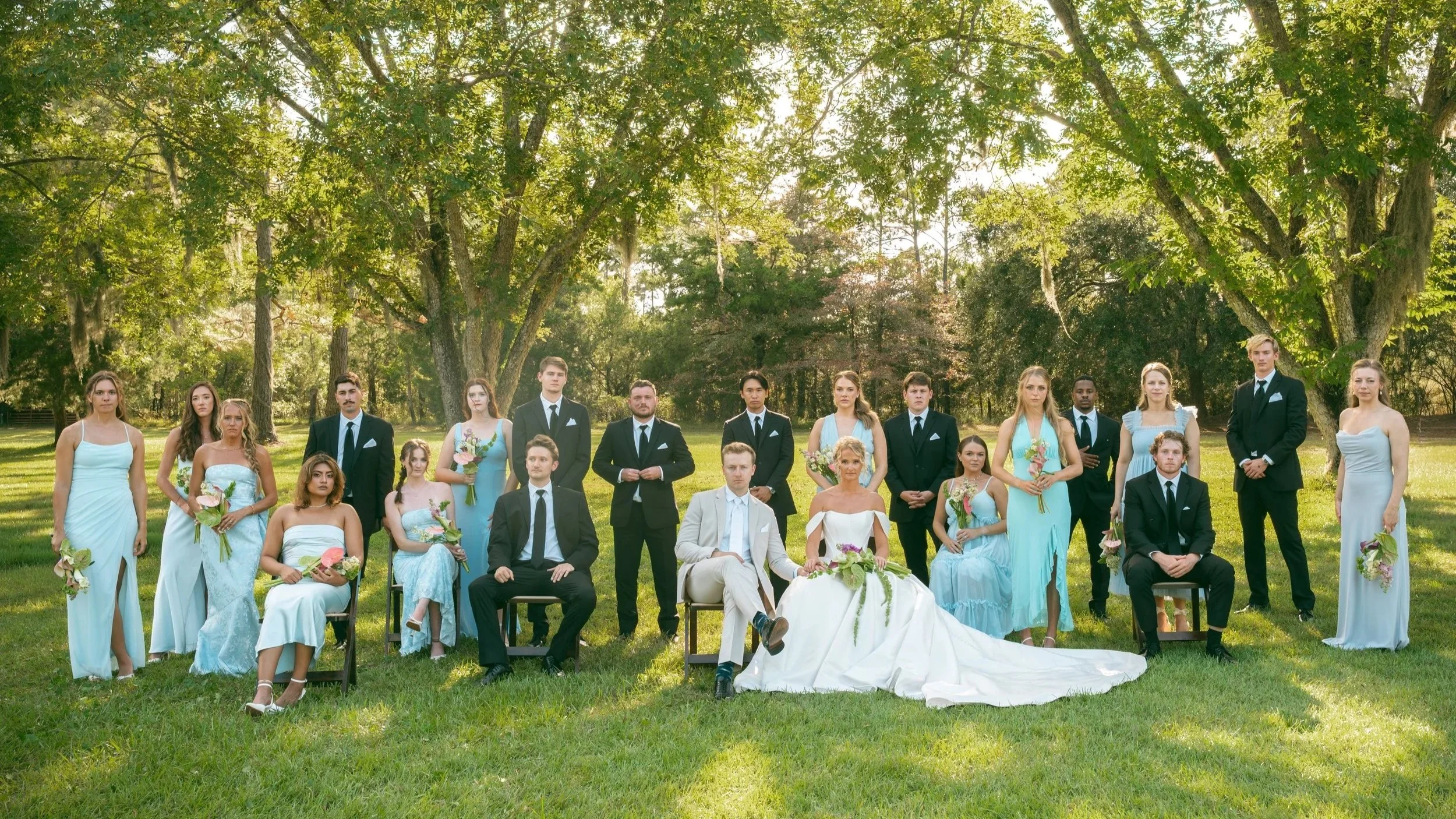 Wedding party outdoors under a large tree, with the bride and groom seated in the center, surrounded by bridesmaids and groomsmen.