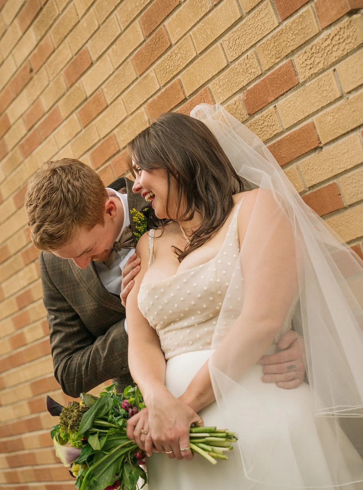 A smiling bride holding a bouquet of flowers, wearing a veil and wedding dress, stands against a brick wall. A groom in a brown checkered suit with a bow tie leans toward her, laughing.