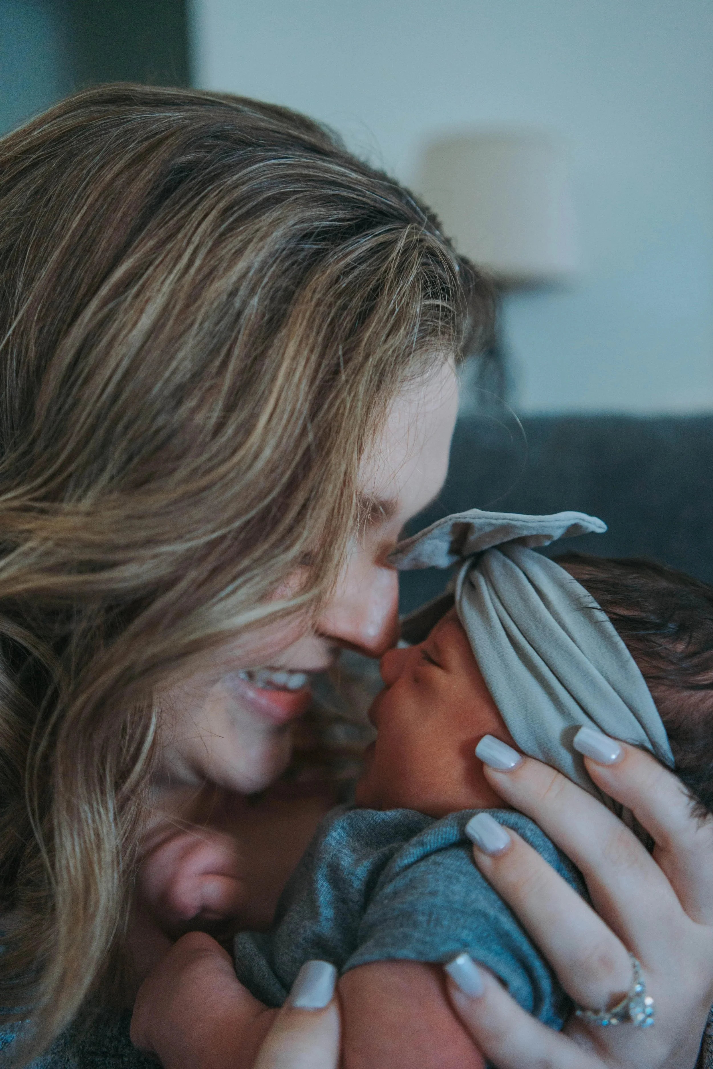 A woman with long, wavy blonde hair gently kissing a young child on the lips. The woman is holding the child's face with both hands, and the child, wearing a gray shirt, is smiling with eyes closed.