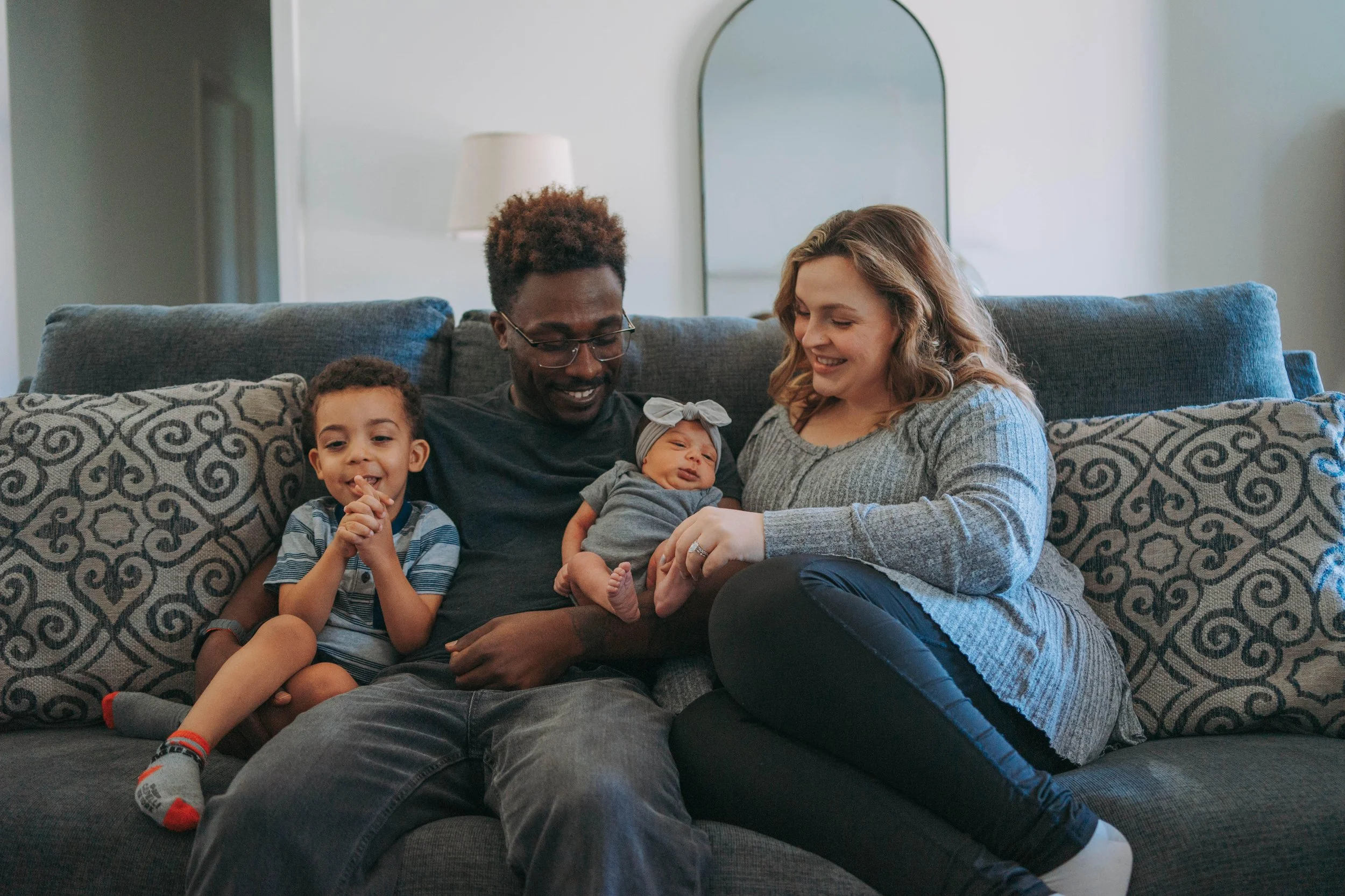 A family of five sitting on a grey couch in a living room, smiling and enjoying time together. The family includes two adults, two children, and a baby.