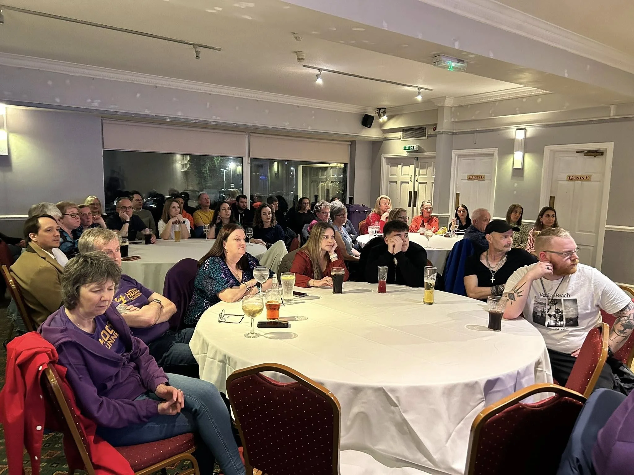 Group of people seated at round tables watching a presentation or speaker in a banquet hall or conference room.