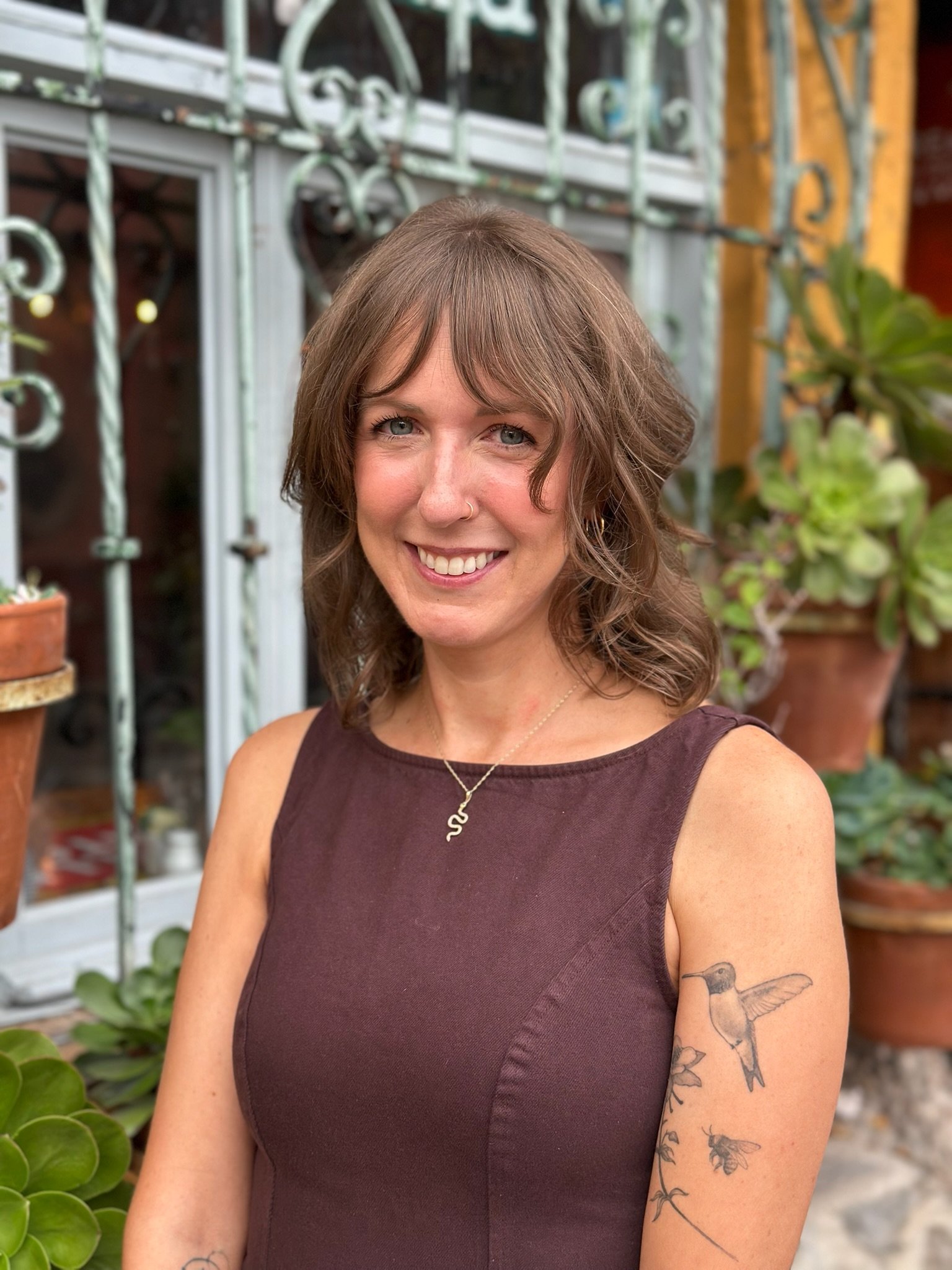 A woman with shoulder-length brown hair, blue eyes, and a nose piercing smiling outdoors in front of a glass greenhouse with potted plants and a brick wall. She is wearing a sleeveless maroon dress, a gold necklace, and has a hummingbird tattoo on her upper arm.