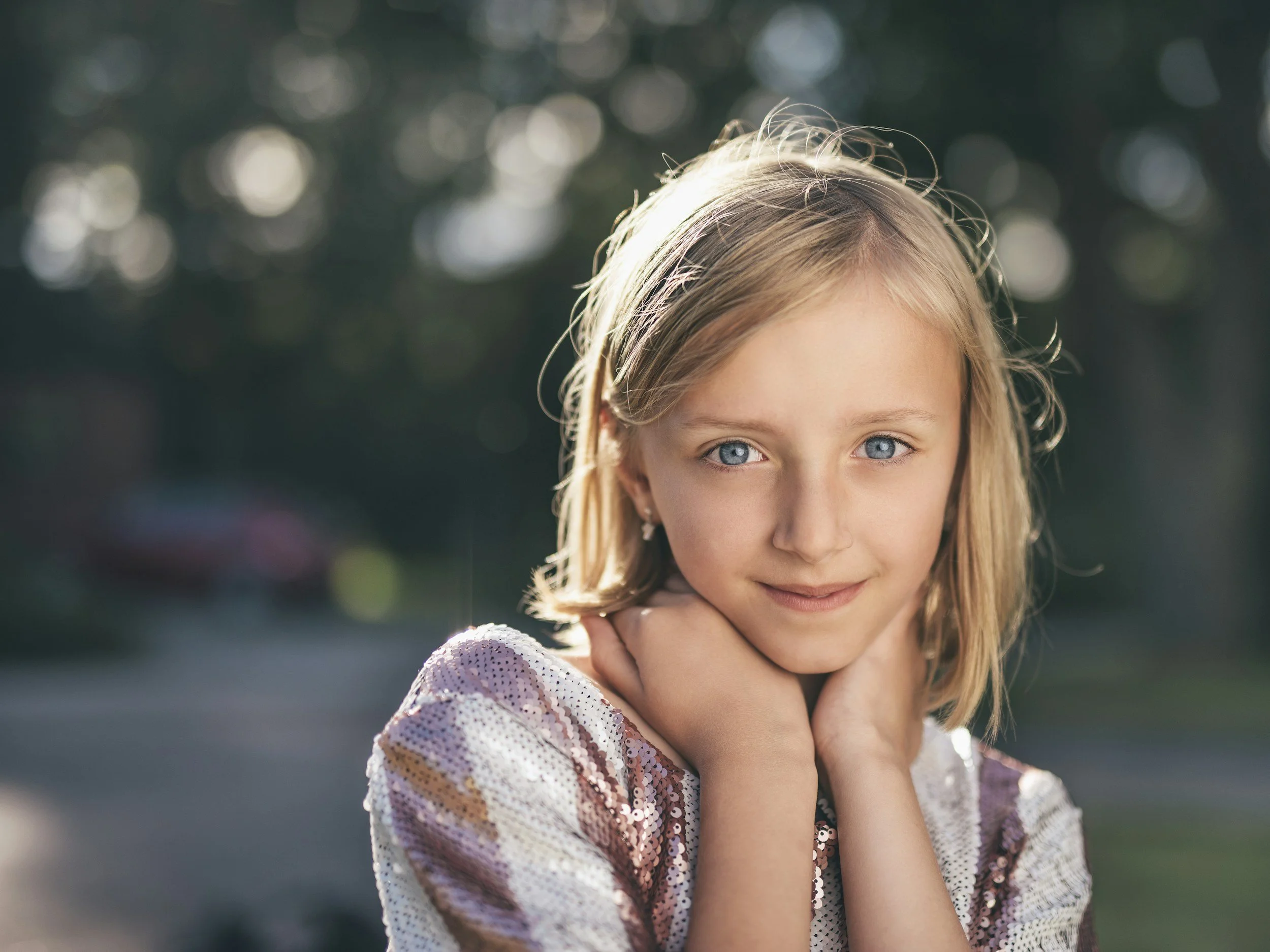 Jeune fille à cheveux blonds et yeux bleus, portant une veste en sequins, posant dehors avec les mains sous le menton, éclairée par la lumière naturelle