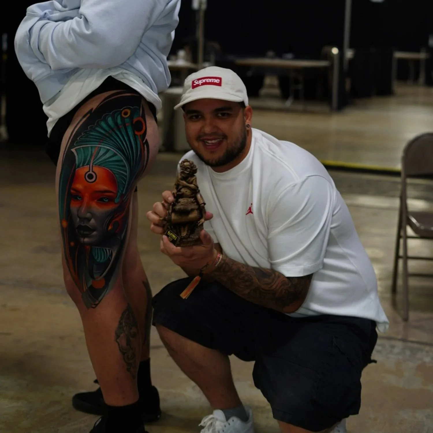 A man wearing a white cap, white T-shirt, and black shorts is crouching and smiling while holding a small bear sculpture. Next to him, a person with a large, colorful tattoo on their leg is standing, partially visible.