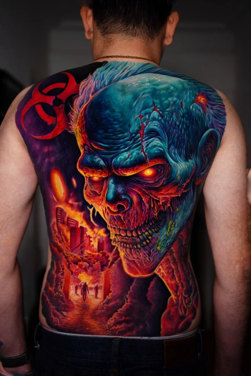 Colorful tattoo of a zombie-like face with glowing eyes on a man's back, incorporating city skyline and fiery elements.