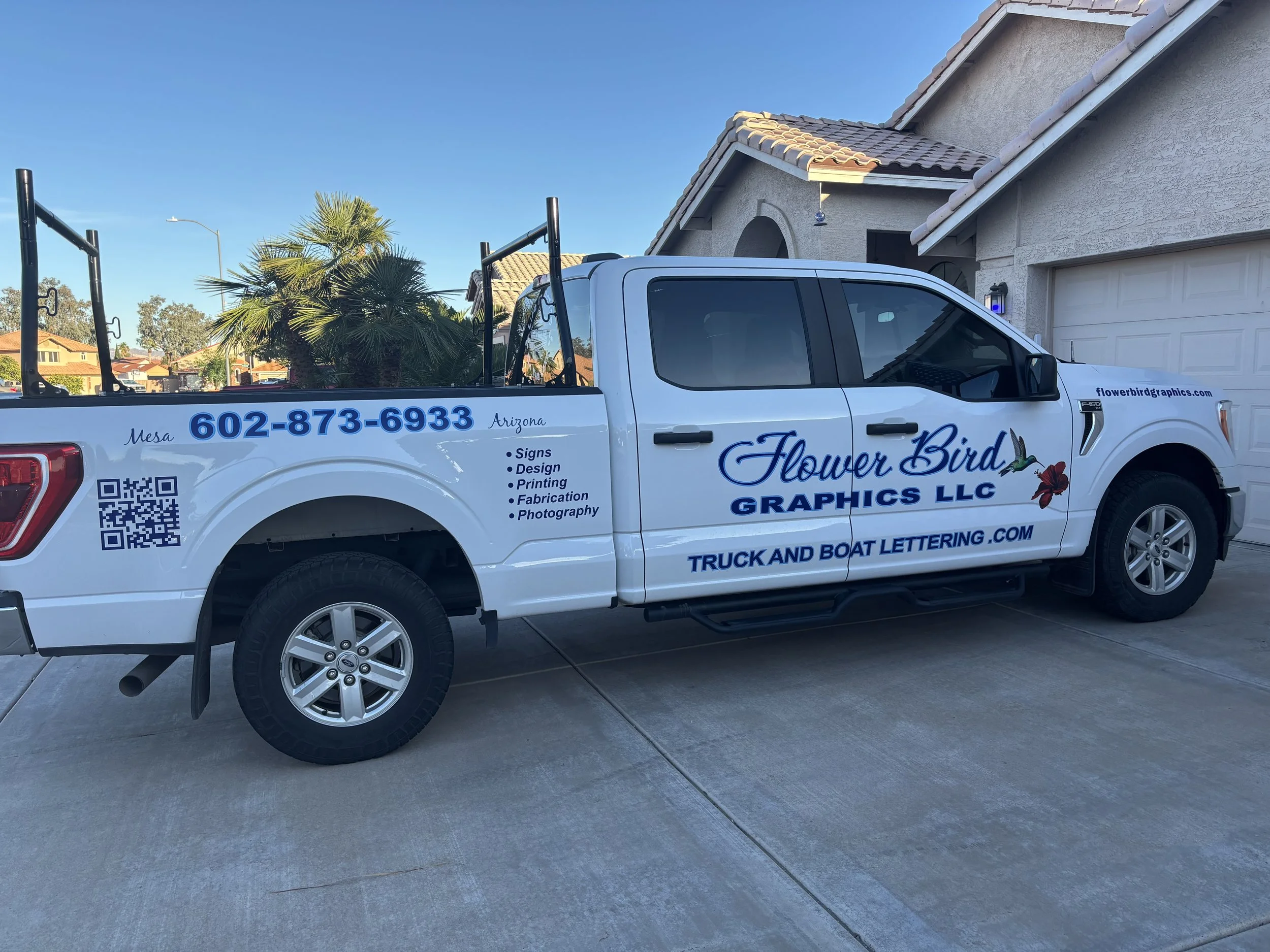 White Ford pickup truck with business branding for Flower Bird Graphics LLC, parked outdoors near a lake, with palm trees and green bushes in the background. The truck displays contact information and services like signs, design, printing, fabrication, and photography.