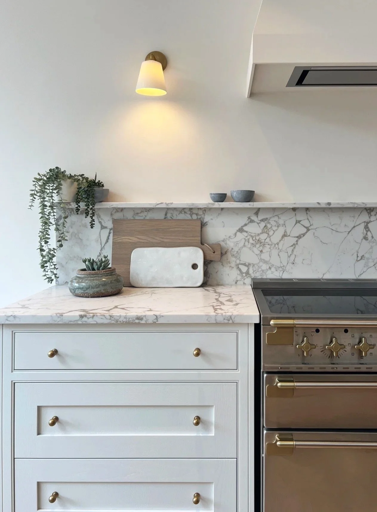 Modern kitchen with white cabinets, brass handles, marble countertop & backsplash, decorative accessories, wall light, and range cooker