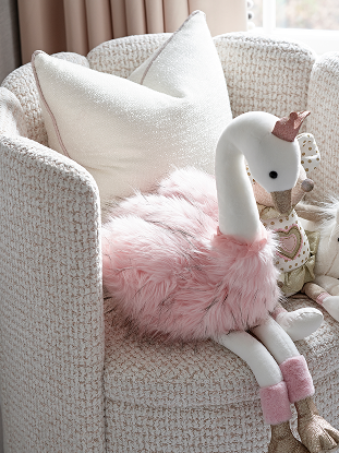 Plush toy swan with pink fluffy body, white neck and head, pink socks, and small crown, sitting on luxury pink and white textured armchair with cushions