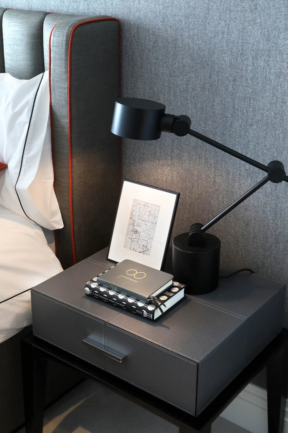 Modern masculine bedroom with gray headboard, white luxury bedding, grey leather bedside table with black desk lamp, framed abstract artwork, and books