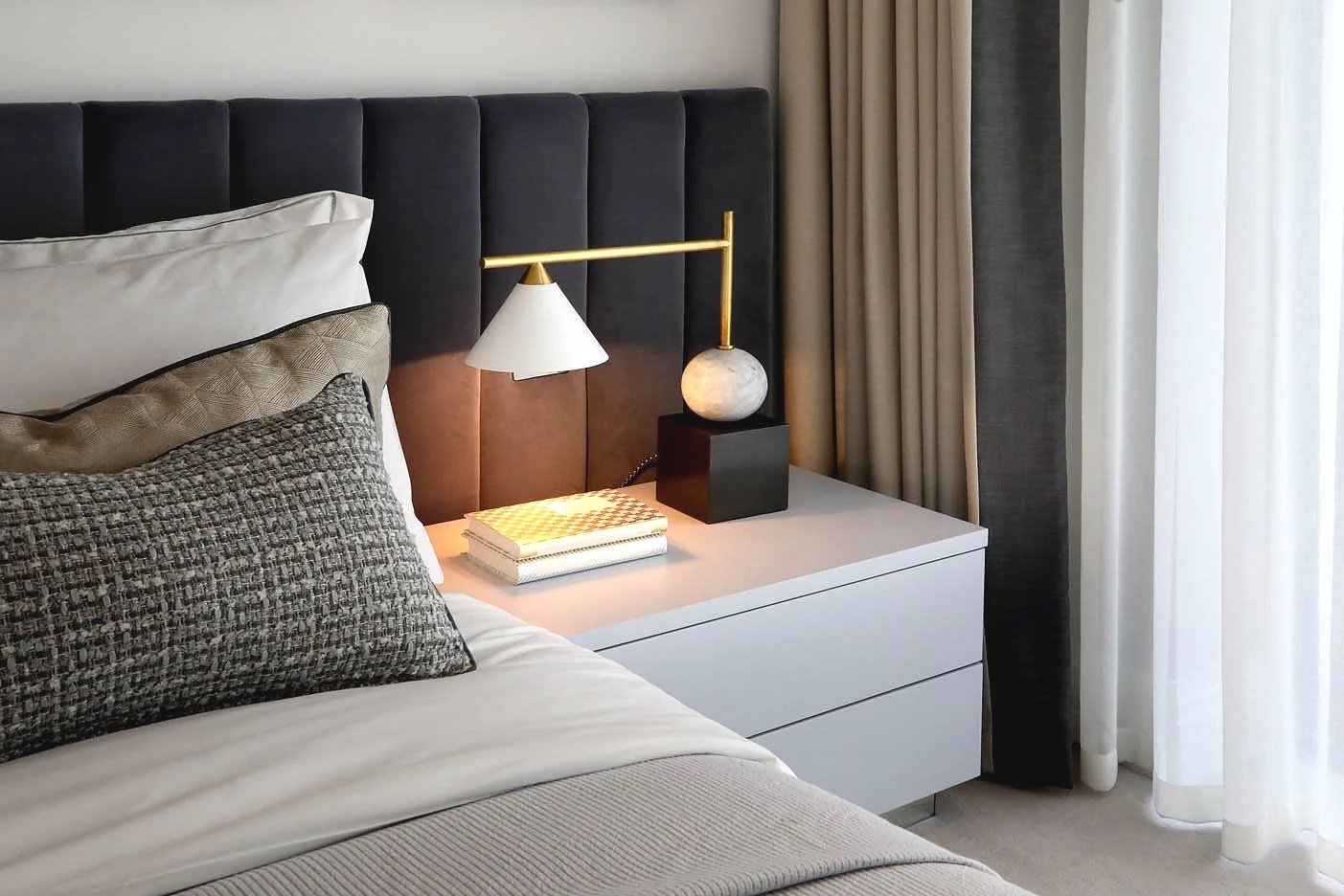 Modern luxury bedroom with bed, layered cushions, bedside table with Kelly Wearstler lamp, books, and window with curtains