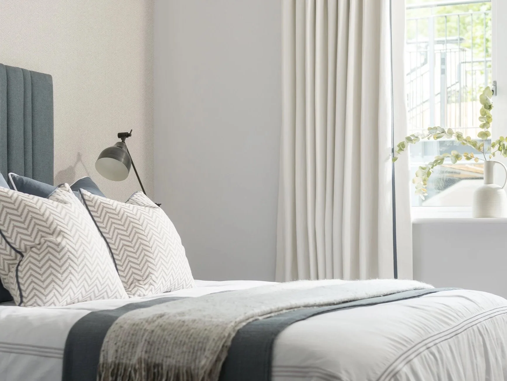 Modern coastal bedroom with white and grey bedding, chevron cushions, black lamp, white curtains, and eucalyptus vase on windowsill.
