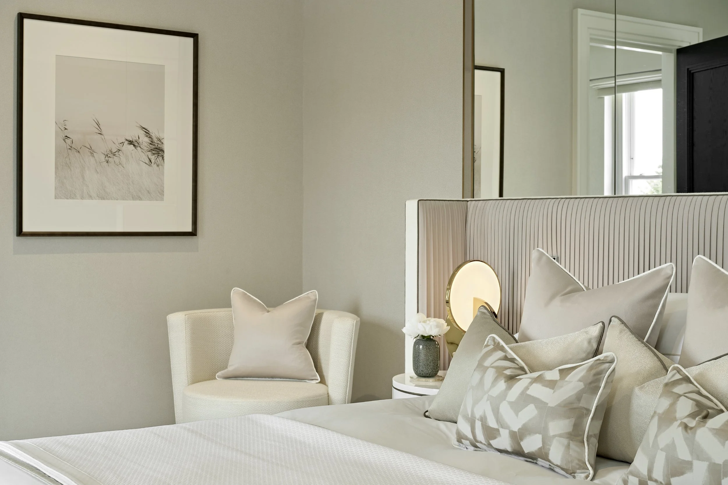 Modern luxury bedroom in soft pinks and neutrals with white bed linen, layered cushions, bedside table, lamp, vase, and artwork