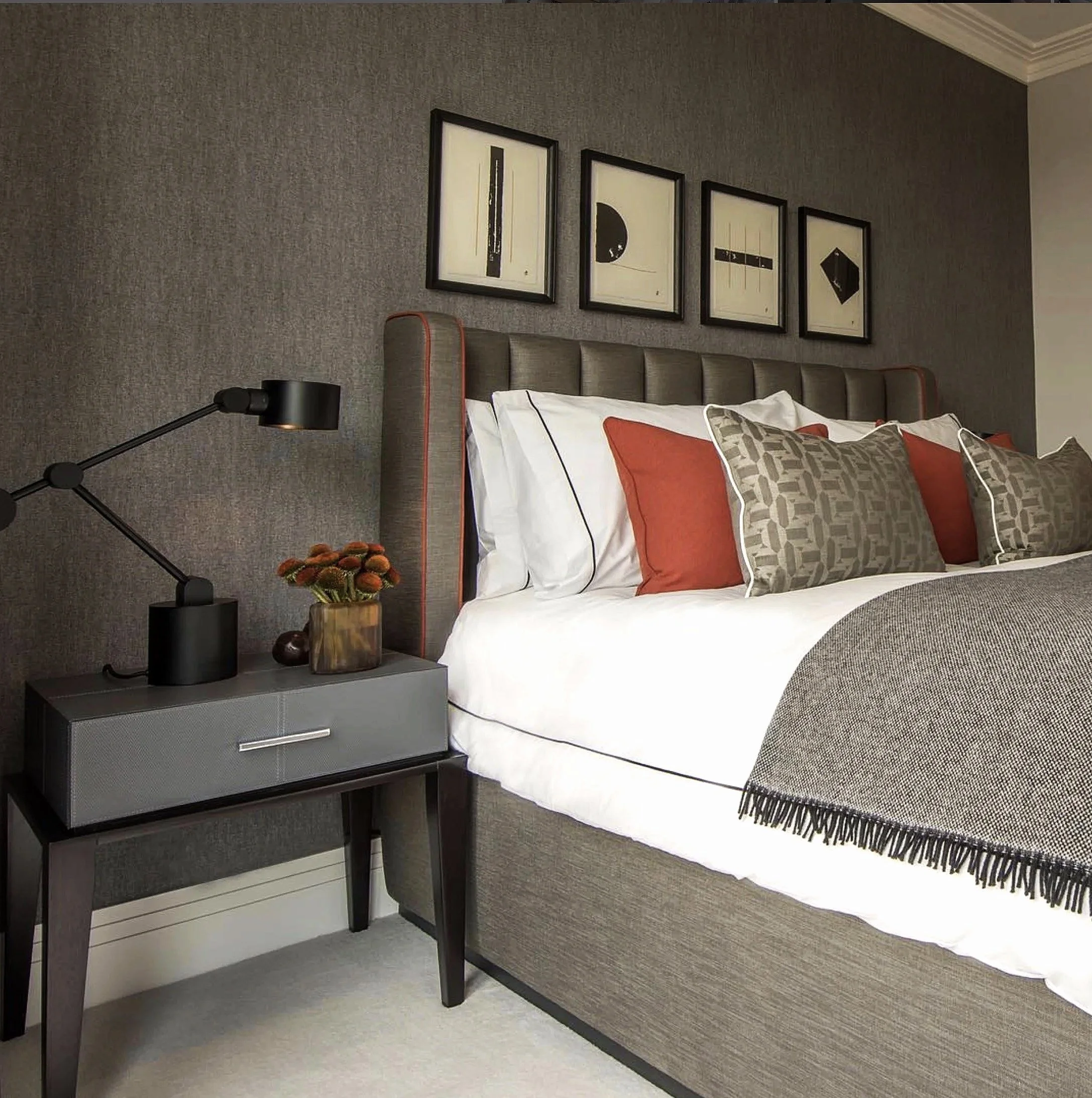 Masculine luxury bedroom with grey bedside table, sculptural flowers, white bedding with red cushion, textured throw, black-and-white abstract artwork, and Tom Dixon lamps