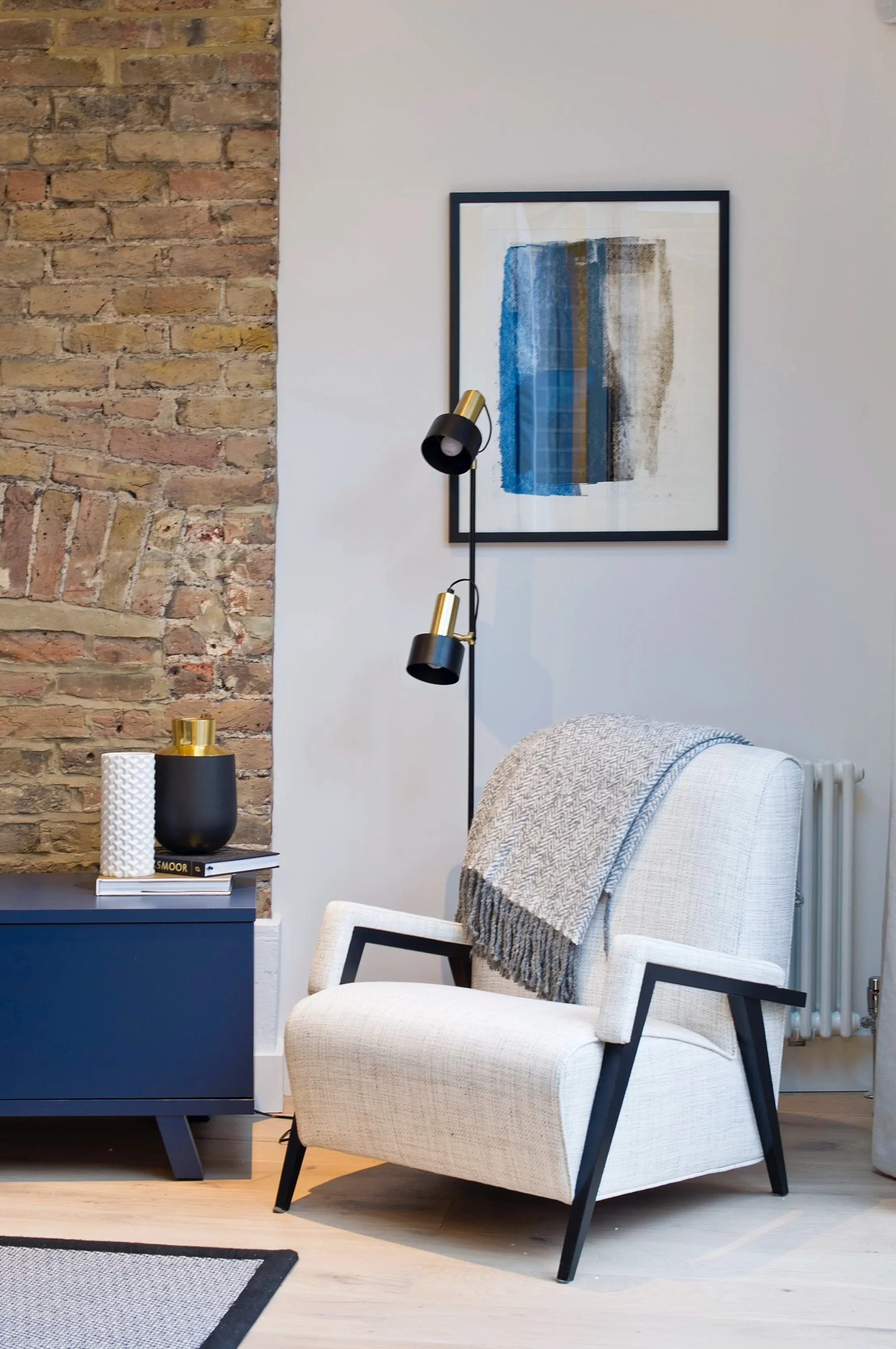 Modern mid-century living room corner featuring a white armchair, blue side table, black and gold lamp, abstract wall art, and exposed brick wall
