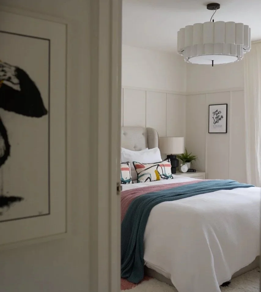 Modern bedroom viewed through an open door featuring white bedding, teal and pink throw, abstract cushions, bedside lamp, and wall panelling