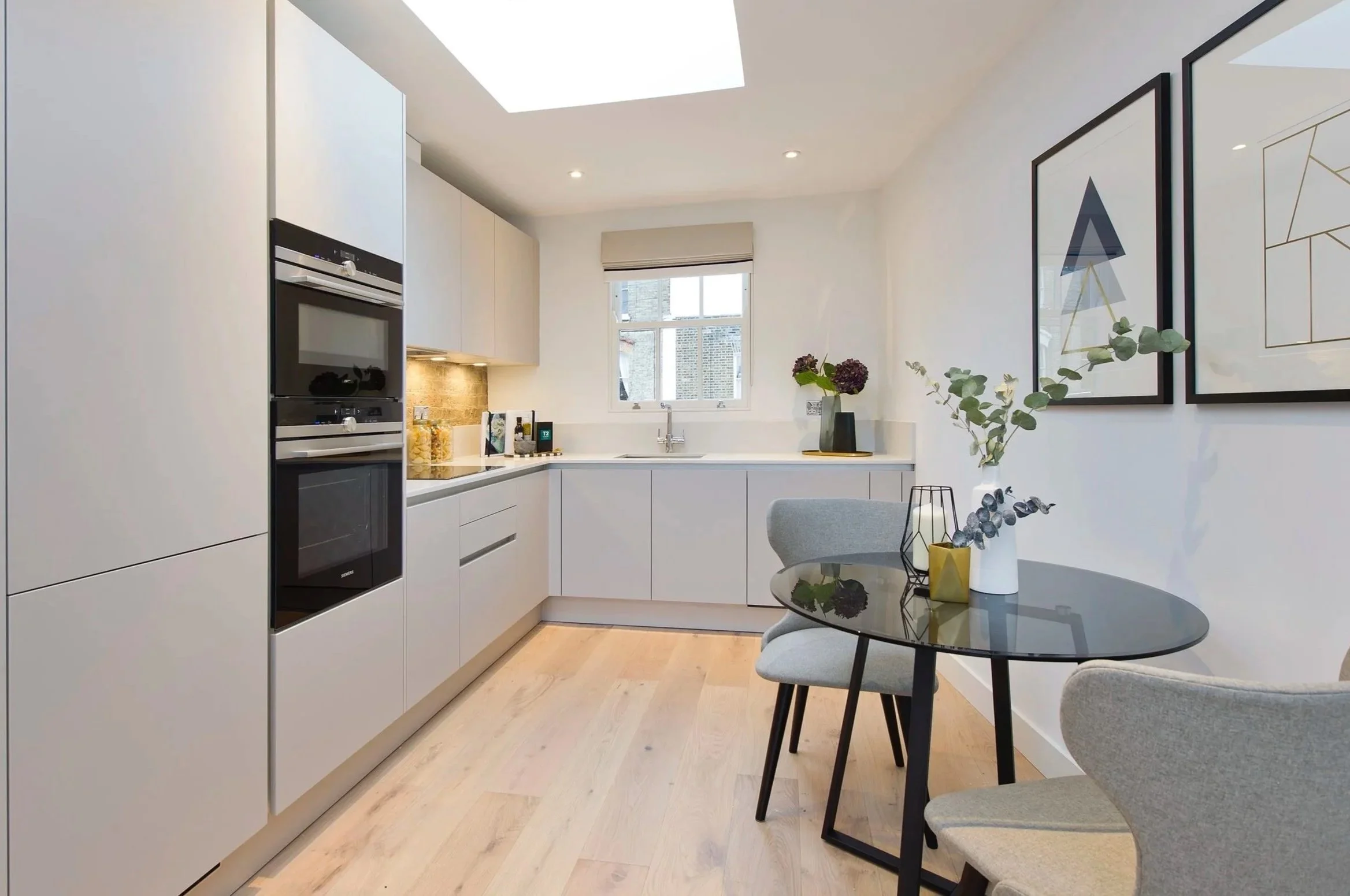 Sleek modern neutral kitchen with round glass dining table, grey chairs, geometric vases, and framed wall art.
