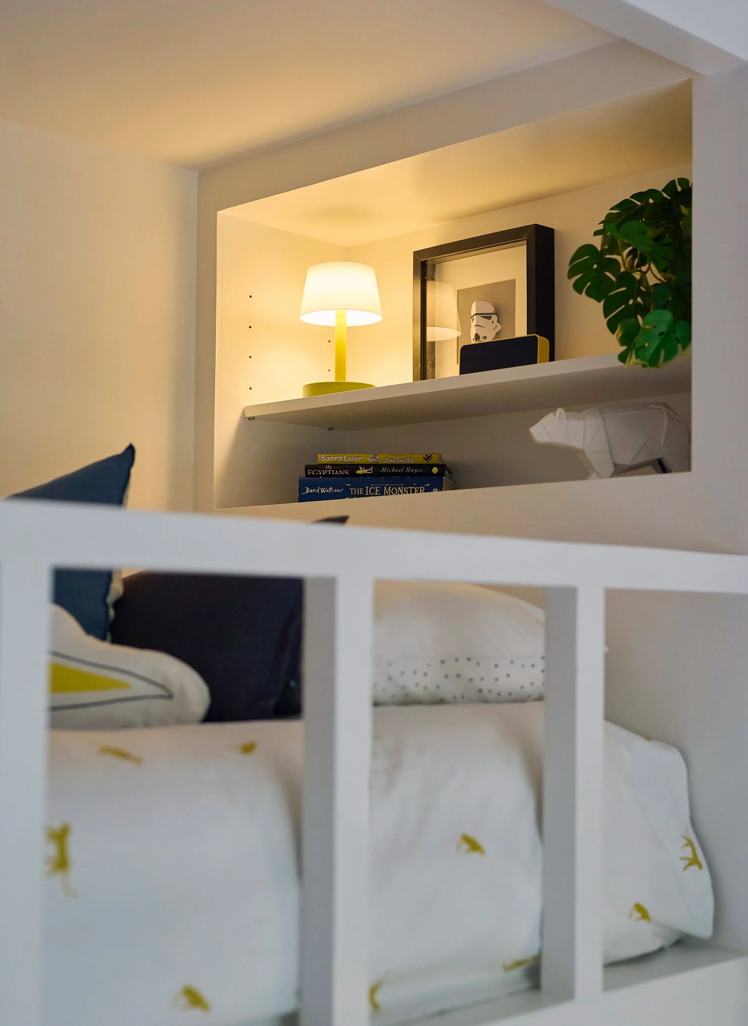 A cozy kids loft bed featuring white bedding and yellow cushion, built-in nook with shelving displaying a lamp, framed pictures, books, and a decorative origami bear.