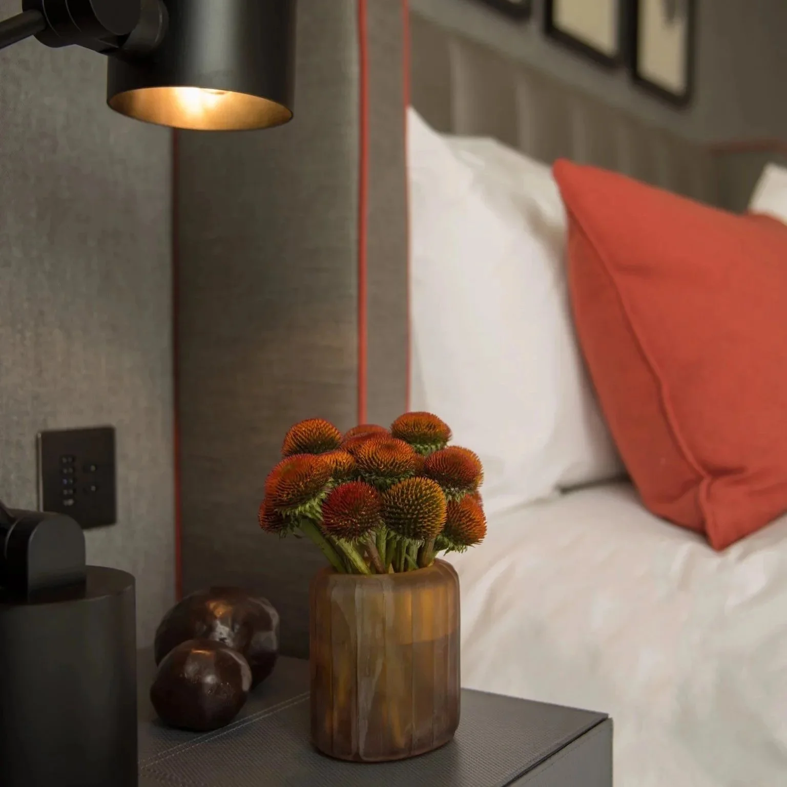 Modern luxury bedroom with grey leather bedside table, sculptural red and green flowers, white bedding with red cushion, Tom Dixon lamps, and framed artwork
