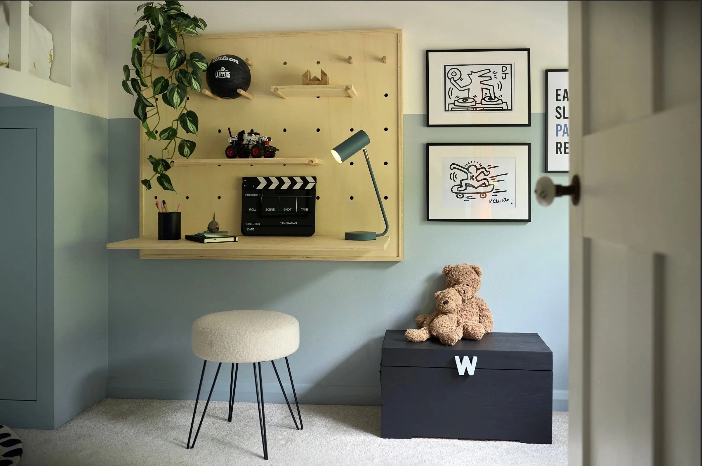 Modern Scandanavian kids’ room corner with toy box, desk and wooden pegboard displaying toys, desk lamp and playful wall art.