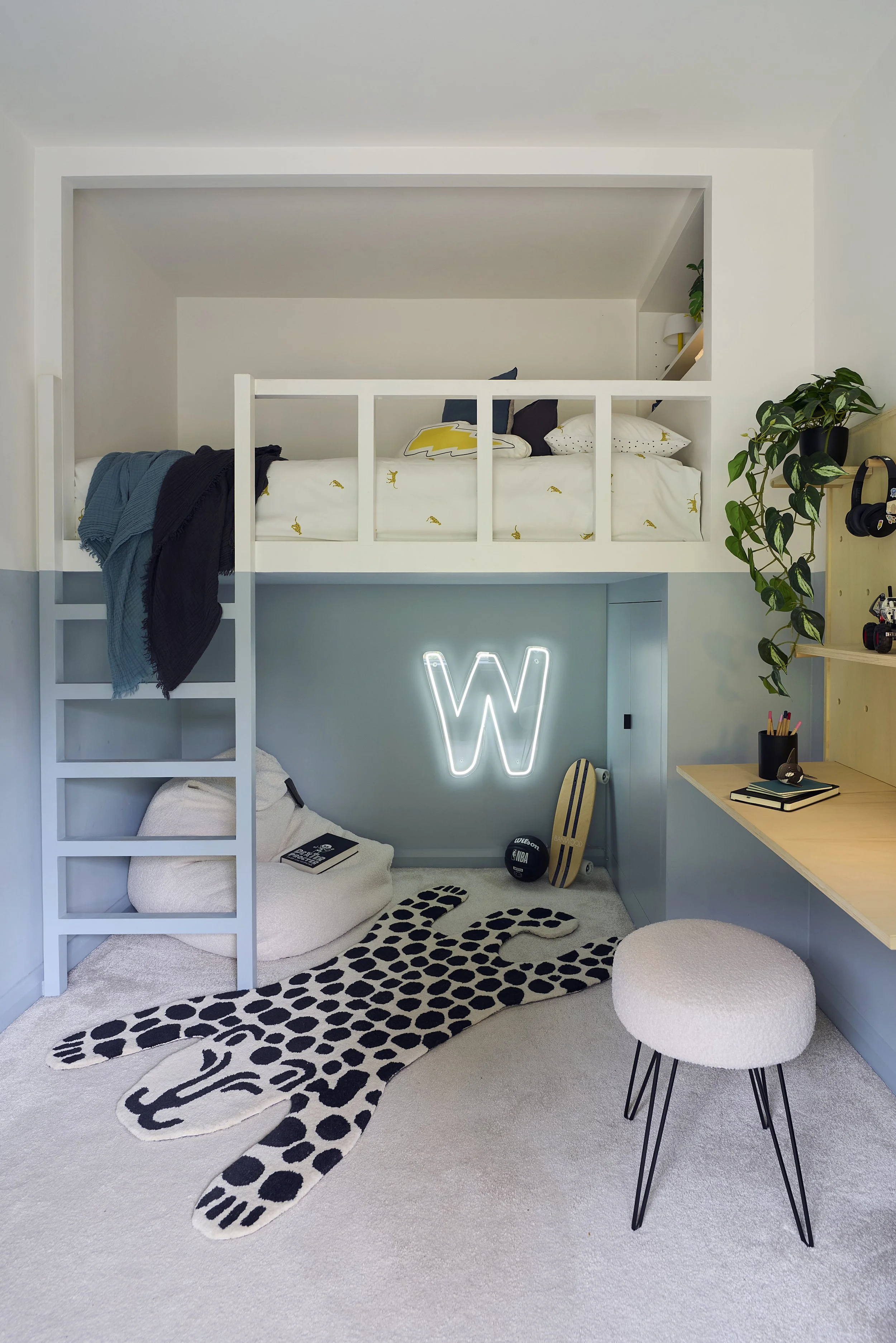Modern kids bedroom with loft bed, white bedding, black and yellow cushions, patterned rug, neon W light, and built-in wooden desk