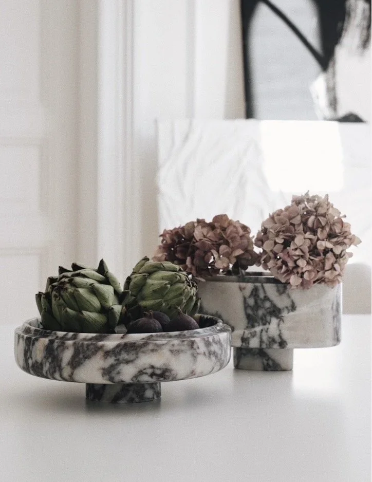 Contemporary marble fruit bowl with artichokes and figs beside a marble vase of dusky pink hydrangeas on a white table.