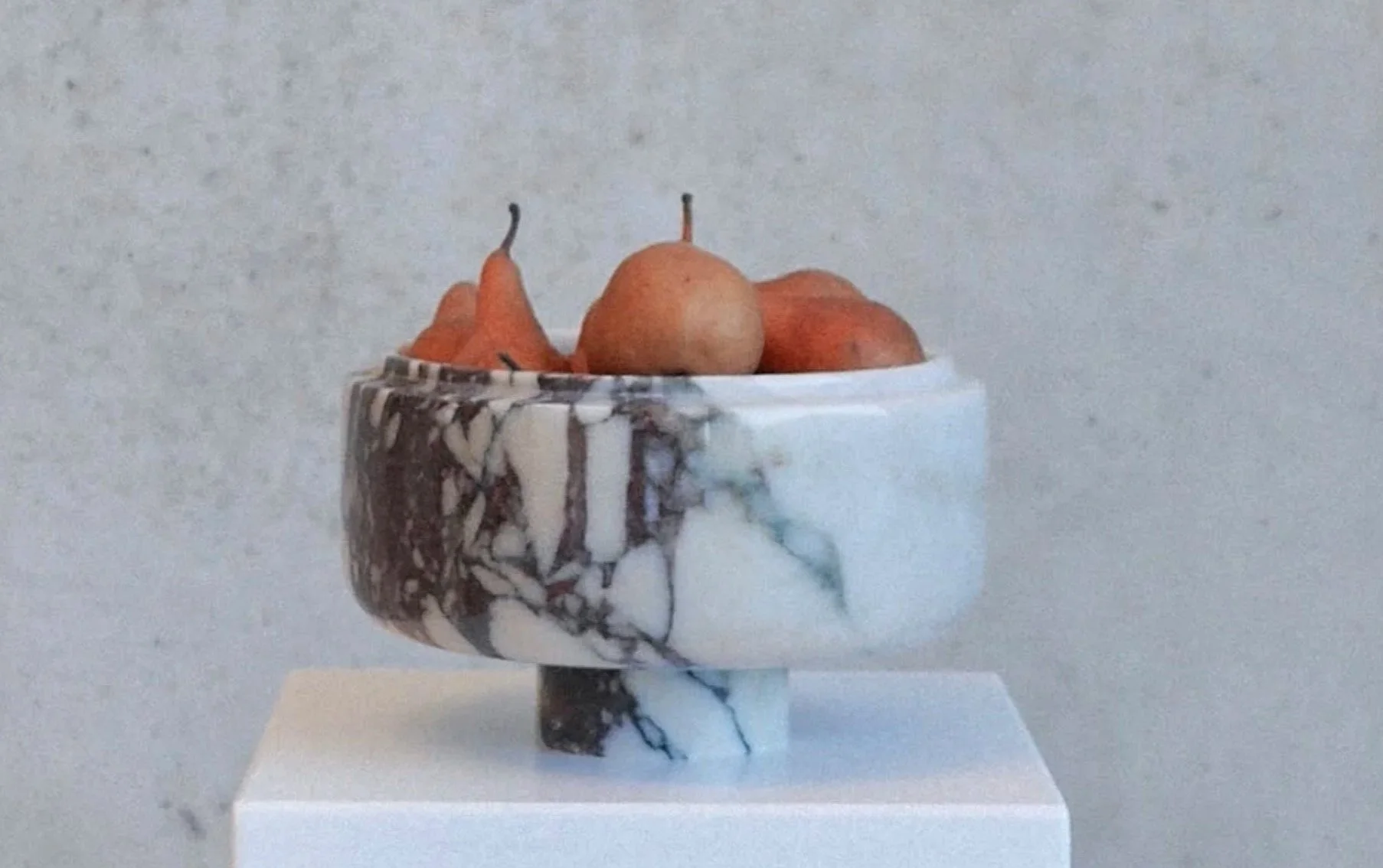 A marble bowl with a black and white marble pattern, filled with pears, placed on a white pedestal against a plain gray wall.