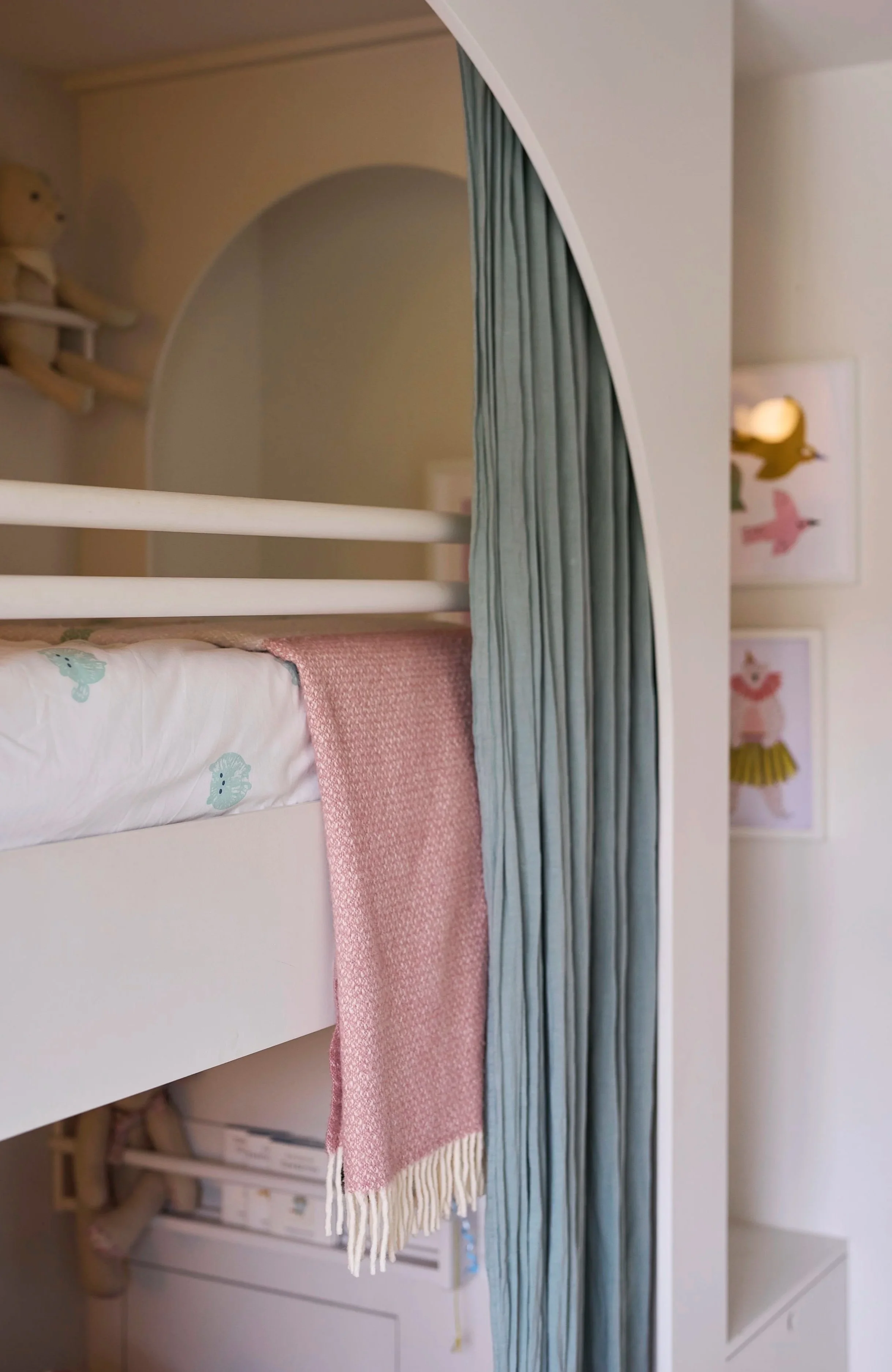Close-up of a child’s arched bunk bed with pink wool blanket, partially drawn green linen curtain, and playful framed pictures on the wall.