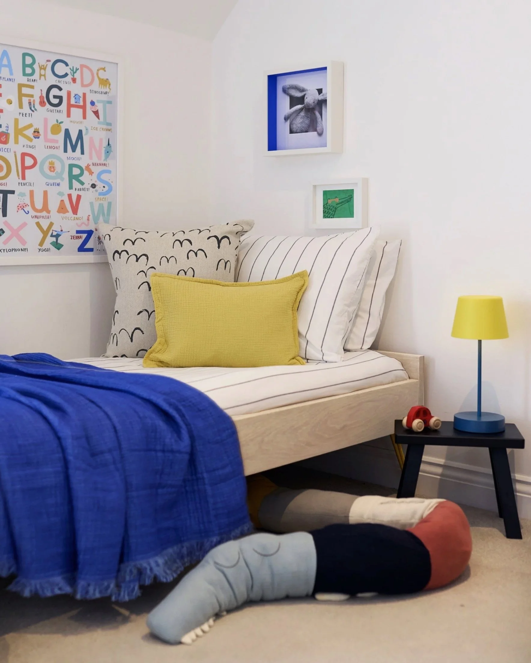 Modern kids’ bedroom with toddler bed, colorful alphabet wall art, animal pictures, nightstand with lamp, toy car, and soft toy.