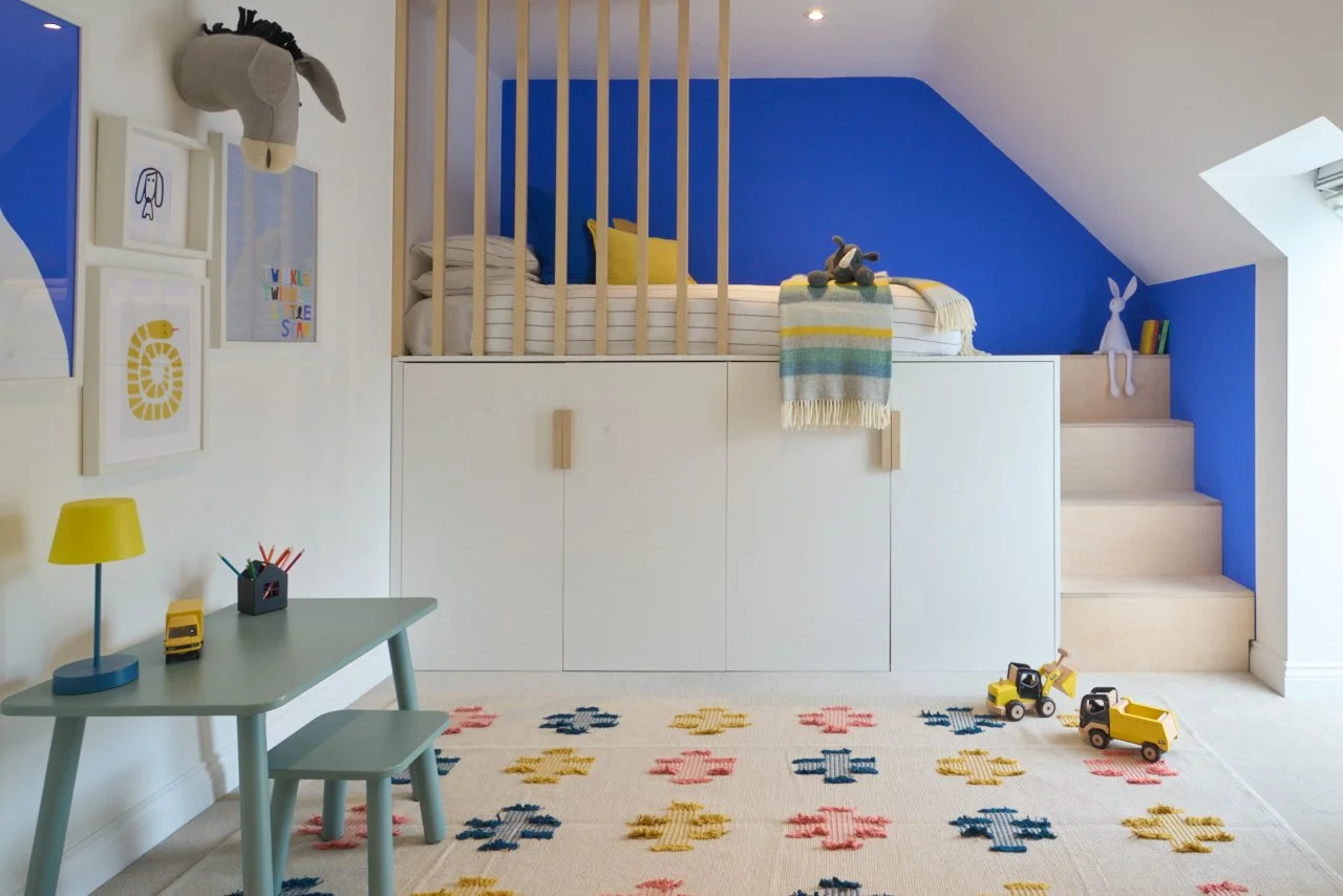 Modern kids’ bedroom with built-in storage bunk bed, electric blue wall, soft toys, bunny lamp, desk, and colorful patterned rug.