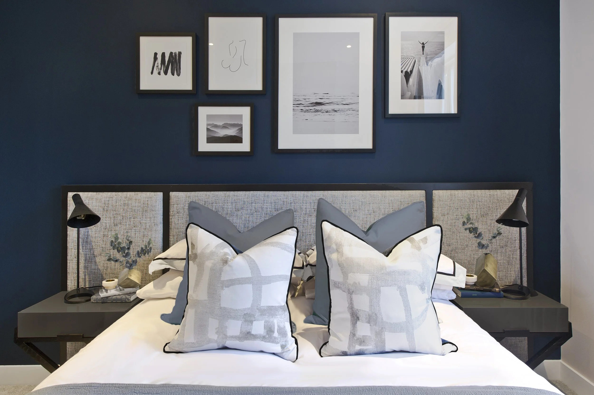 Modern luxury bedroom featuring wide textured headboard, abstract cushions, black lamps, and dark blue wall with framed prints.