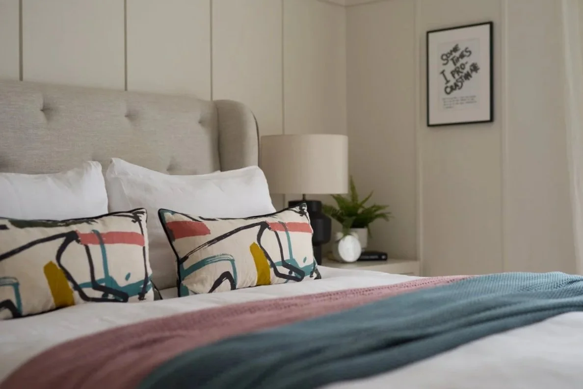 Modern panelled bedroom with beige upholstered headboard, white bedding, abstract cushions, textured blanket, bedside table with lamp, plant, and framed quote