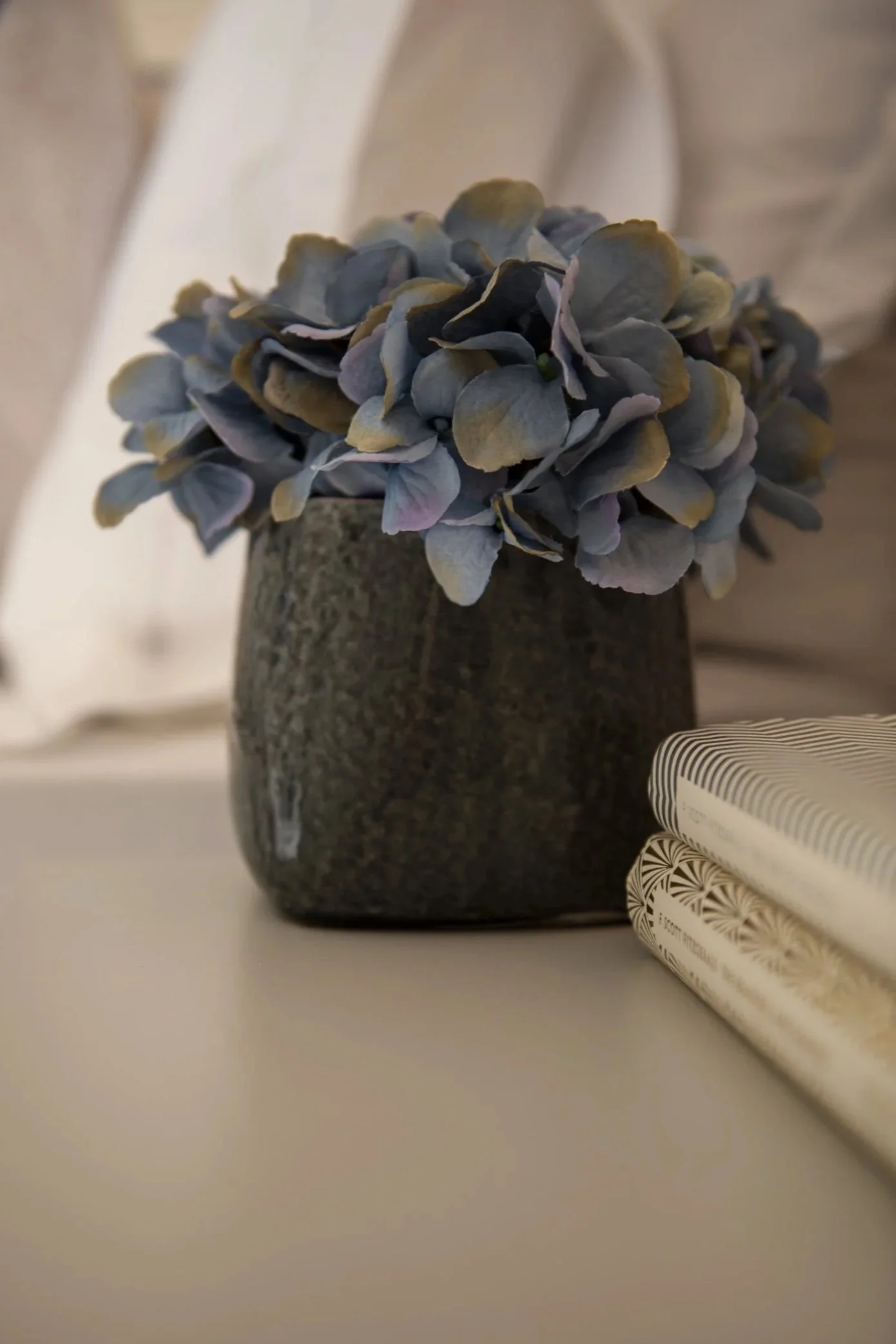 Modern decorative accessories on bedside table: handblown grey glass vase with purple and blue hydrangeas, next to two patterned books.