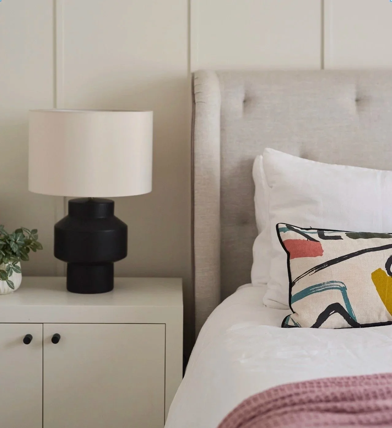 Scandinavian bedroom with light grey upholstered headboard, white bedding, colorful abstract cushion, white bedside table with black knobs, black lamp with white shade, and small potted plant.