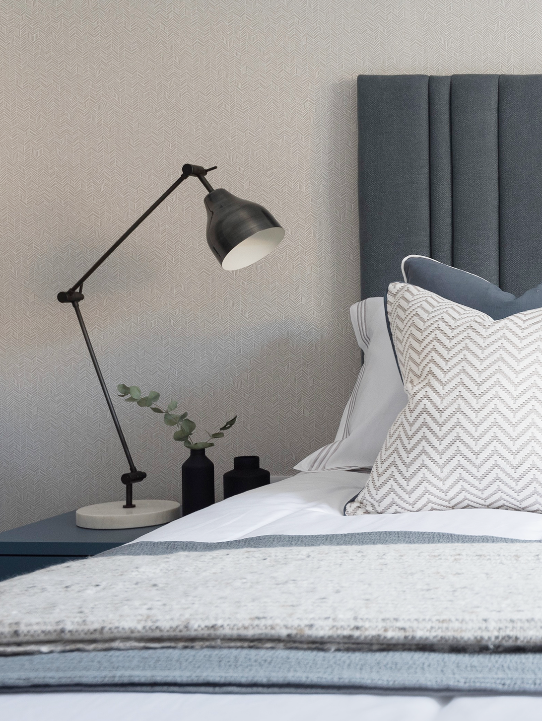 Contemporary bedroom with white bedding, geometric cushions, gray bolstered headboard, herringbone wallpaper, lamp, and decorative vases.