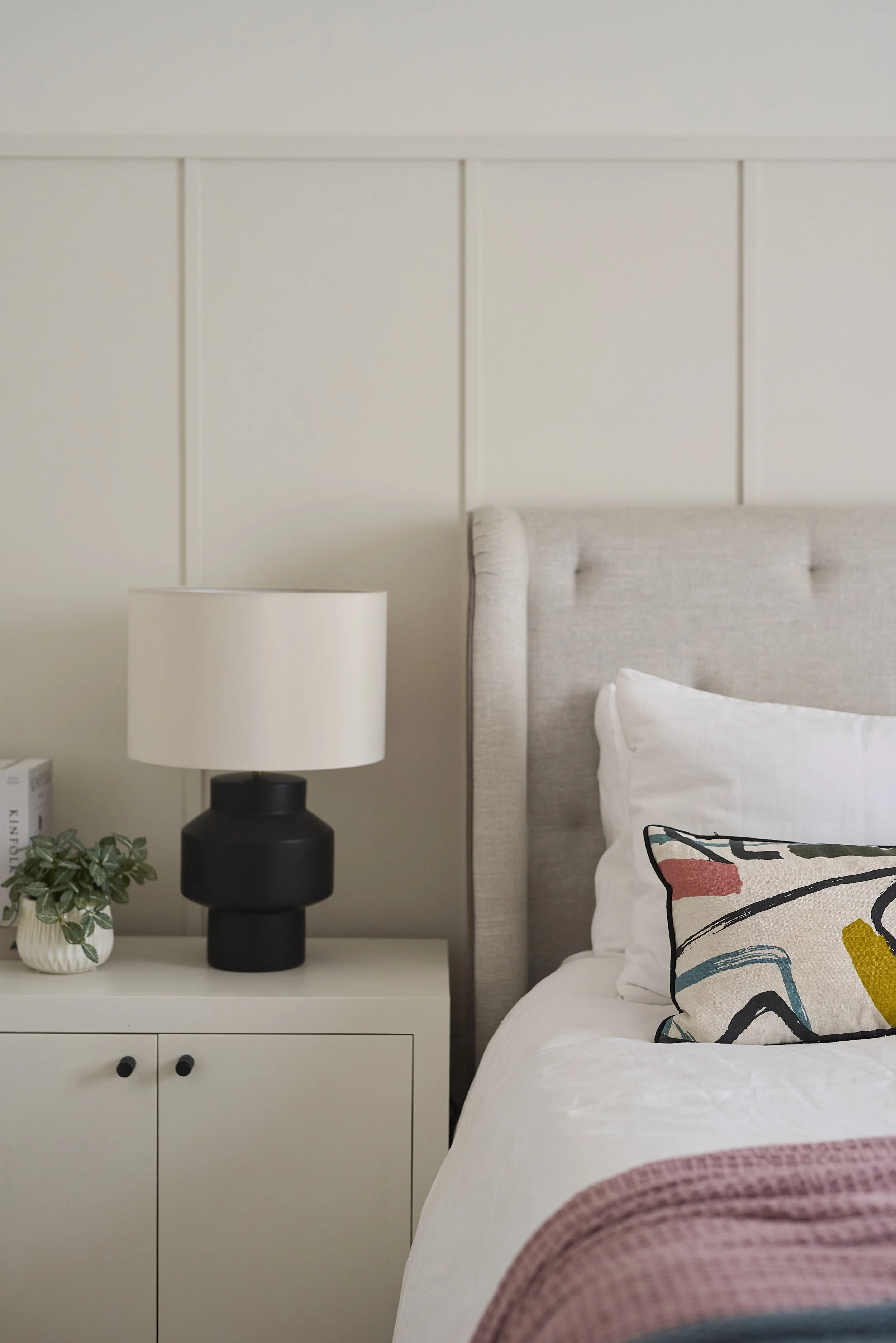 Modern luxury bedroom with beige upholstered headboard, vertical wall panelling, white bedding, colourful abstract cushions, and pink throw