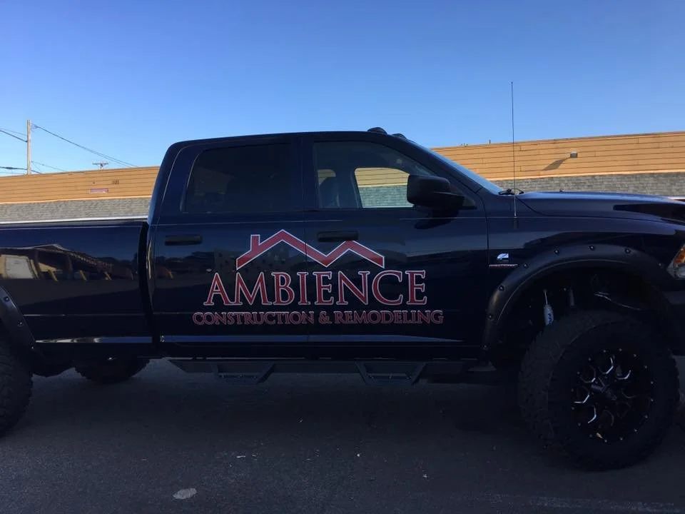 A black pickup truck with the logo of 'Ambience Construction & Remodeling' on the side, parked outside against a building with a blue sky background.