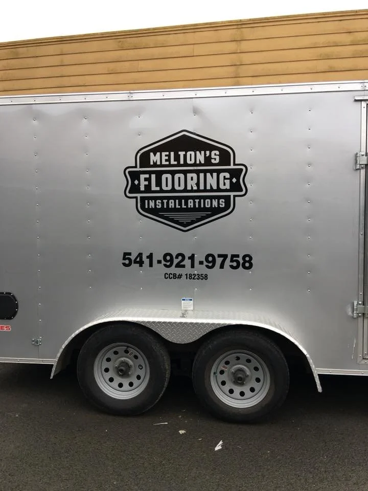 A silver trailer for Melton's Flooring Installations with a phone number and company logo, parked on a dark surface with a wooden wall in the background.
