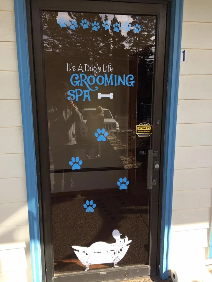 Glass door with blue paw prints, white text saying 'It's A Dog's Life GROOMING SPA' with a white dog in a bathtub graphic, and a yellow 'Stanley' security sticker.