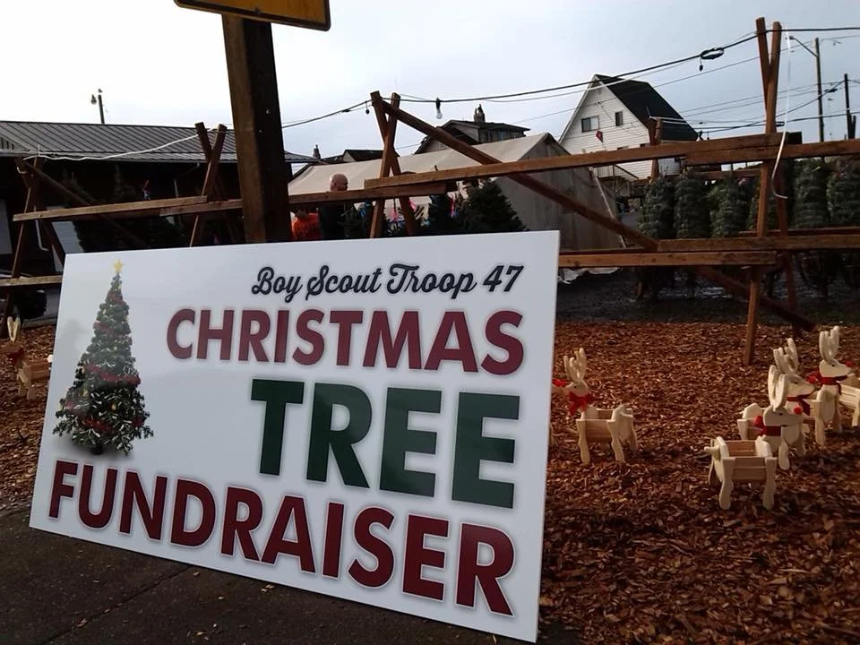 Sign for Boy Scout Troop 47 Christmas Tree Fundraiser with a decorated Christmas tree graphic, placed in front of a small Christmas tree display with miniature reindeer figures and wood scaffolding in the background.