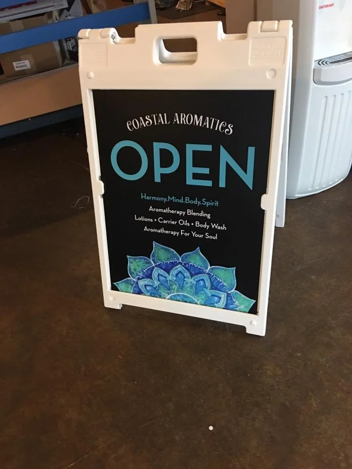 A sidewalk sign for Coastal Aromatics advertises that the store is open, offering aromatherapy products like lotions, carrier oils, and body wash, emphasizing harmony, mind, body, and spirit.