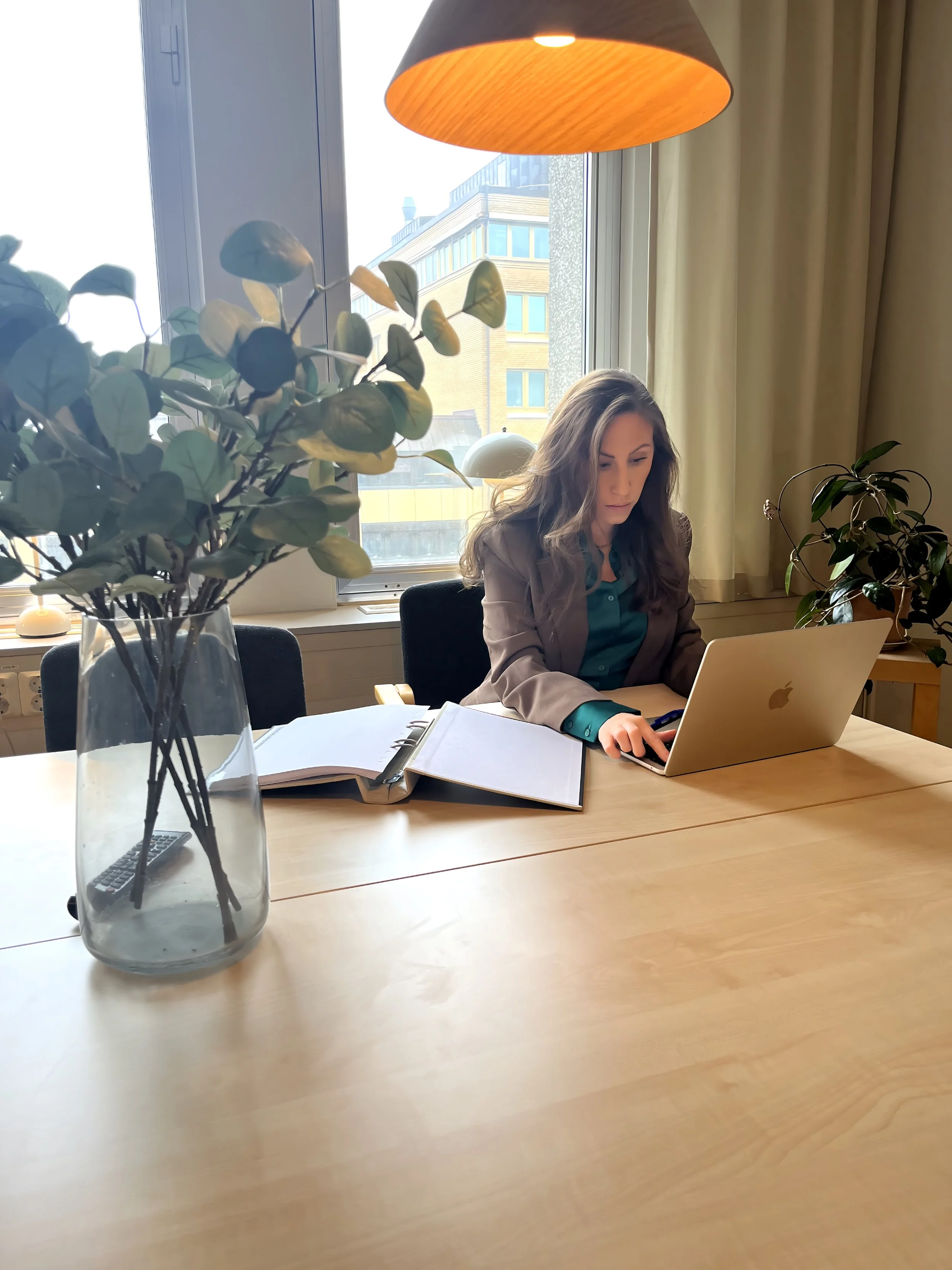 En kvinna som sitter vid ett bord med en laptop, ett anteckningsblock och en växt, i ett rum med stort fönster och gardiner, samt ett taklampor ovanför.