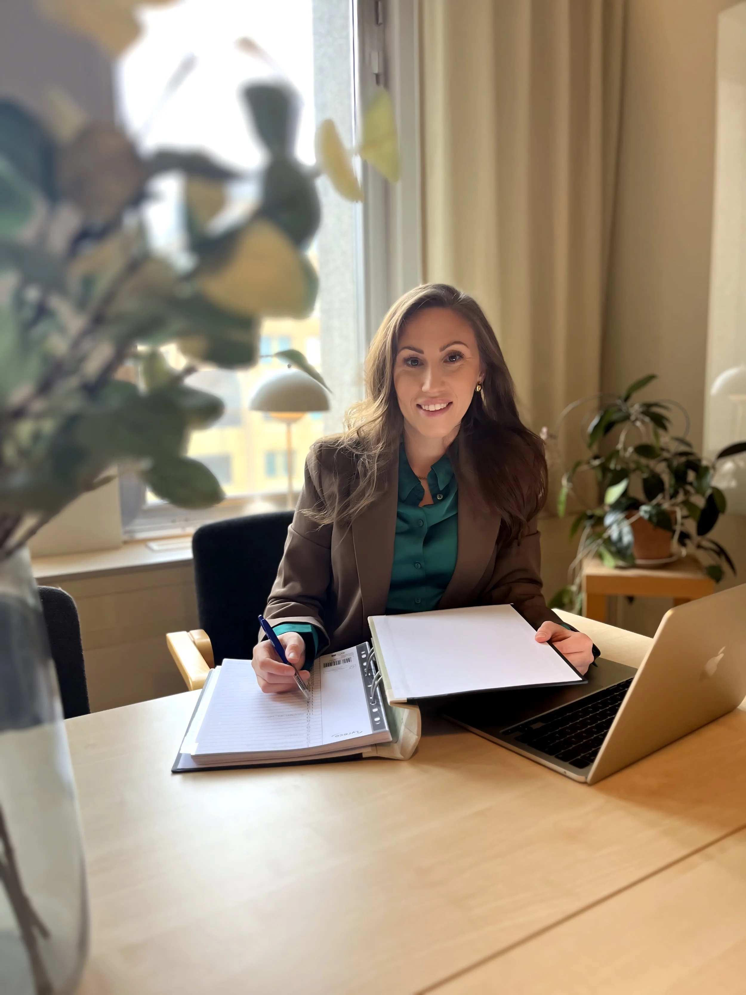 En kvinna som sitter vid ett bord med en öppen laptop och anteckningsböcker, på ett kontor med stora fönster och växter.